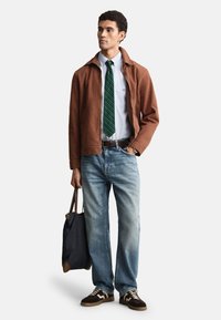 Braune Jacke über einem hellblauen Hemd, grün gestreifte Krawatte, hellblaue Jeans und braune Turnschuhe. Eine marineblaue Einkaufstasche wird in einer Hand gehalten.