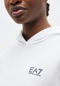 Gros plan d'une personne portant un sweat à capuche blanc avec un logo EA7 Emporio Armani bleu marine sur la poitrine, montrant la partie inférieure de son visage et son cou.