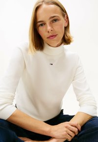 Junge Frau mit blonden Haaren, die einen weißen Rollkragenpullover mit einem kleinen Logo trägt, sitzt im Schneidersitz vor einem schlichten Hintergrund.