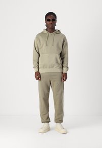 Olivengrønt fleece-hoodie med en frontlomme, parret med matchende sweatpants. Båret med lyse sneakers og sorte solbriller.