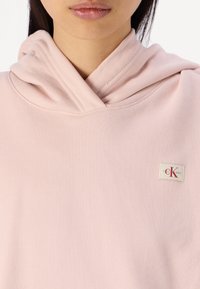 Hoodie rosa chiaro con una texture morbida e un cappuccio a doppio strato. Presenta un piccolo etichetta del marchio con lettering rosso sul davanti.