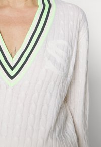 Weißer Kabelstrickpullover mit tiefem V-Ausschnitt, ausgestattet mit einem schwarz und neongrünen gestreiften Kragen und einem dezenten Logo-Detail auf der Brust.