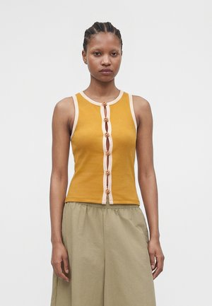 Femme aux cheveux tressés portant un haut sans manches couleur moutarde avec des bordures beiges et des boutons décoratifs, associée à un pantalon beige et ample.