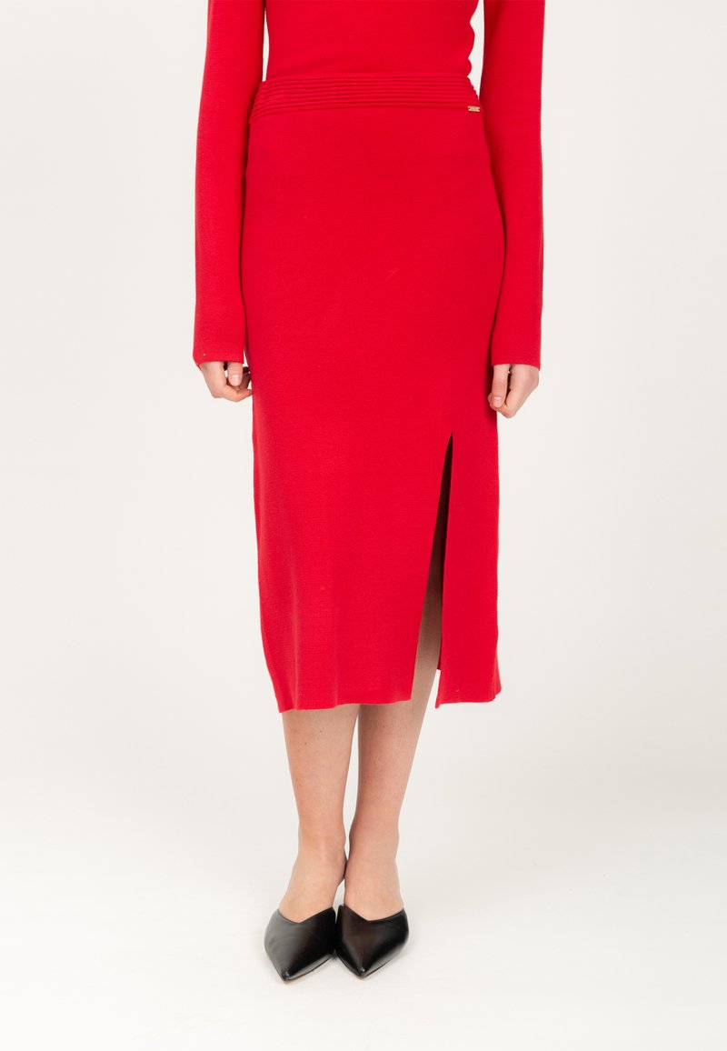 Roter Strick-Midirock mit seitlichem Schlitz und strukturiertem Taillendetail. Kombiniert mit schwarzen spitzen Ballerinas auf einem neutralen Hintergrund.