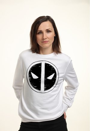 Donna con capelli castani fino alle spalle che indossa una felpa bianca con un grande logo Deadpool in bianco e nero, in piedi con una mano in tasca.