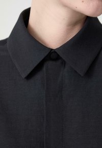 Černá košile s límečkem a knoflíkovým zapínáním, vyrobená z hladkého materiálu a se strukturovaným designem, zaměřující se na čistý, minimalistický vzhled.