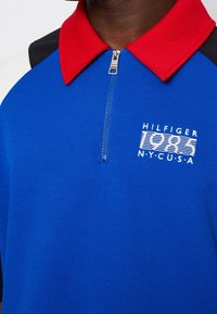Polaire bleu avec col rouge et accents noirs sur les épaules, comportant une fermeture éclair à l'avant et un logo brodé avec "HILFIGER 1985 NY.C.U.S.A."