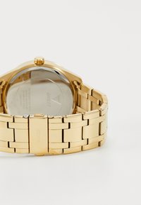 Guld rustfrit stål ur med et link armbånd. Rund urkasse med en gennemsigtig bagside og en fremtrædende krone på siden.