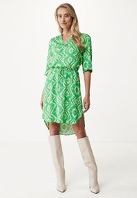 Grünes gemustertes Hemdkleid mit einem Taillengürtel, langen Ärmeln und einem Vorderteil, das kürzer ist als das Rückenteil. Kombiniert mit knielangen weißen Stiefeln. Glatte Textur.