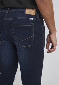 Vaqueros azul oscuro de denim, con una etiqueta de cuero marrón que dice "11 PROJECT" y costuras amarillas en los bolsillos.