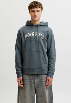 Ung mand iført en blå Jack & Jones hættetrøje med frontlomme og falmede grå jeans, stående mod en ensfarvet lys baggrund.