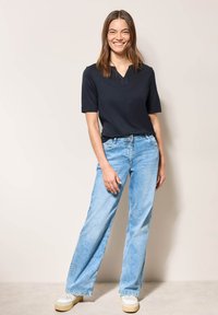 Marineblå kortærmet top med V-hals, parret med lyseblå vide jeans og hvide sneakers, sat mod en neutral baggrund.