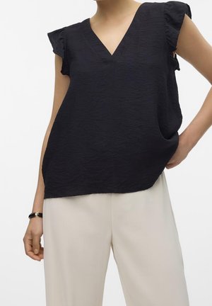 Vrouw draagt een zwarte gestructureerde mouwloze V-hals top met gerimpelde schouders en lichtbeige broek, hand rustend op de heup.
