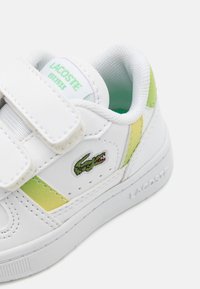 Baskets en cuir blanc avec des accents verts et dorés, dotées de sangles Velcro ajustables et d'un logo crocodile brodé.