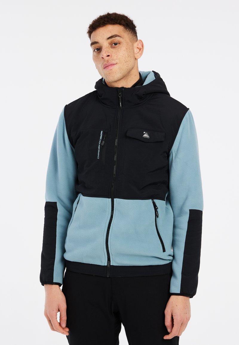 Capuchonjack met een zwart bovenstuk en een lichtblauw fleece onderstuk. Voorzien van ritszakken en verstelbare manchetten voor een goede pasvorm.