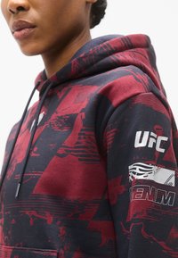 Sweat à capuche bleu marine et rouge, avec un motif graphique, des cordons de serrage et des logos imprimés sur la manche. Tissu doux en mélange de coton.