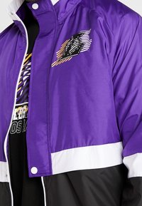 Lila vindjacka med vita och svarta accenter, med en grafisk logotyp på bröstet. Slät, lättviktsmaterial med dragkedja.