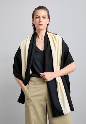 Mujer con el pelo peinado hacia atrás, que lleva una camisa negra, pantalones beige y una bufanda negra y crema colocada sobre los hombros frente a un fondo liso.