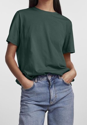 Mujer con camiseta de manga corta verde oscuro ligeramente metida en jeans de tiro alto azul claro, con las manos en los bolsillos.