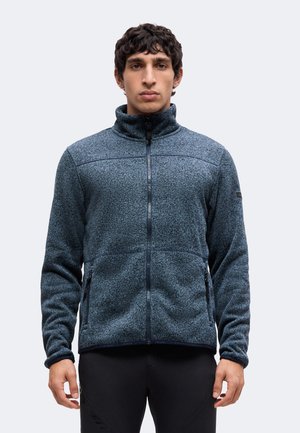 Homme portant une veste polaire bleue zippée avec un col montant et un pantalon noir, debout devant un fond clair uni.