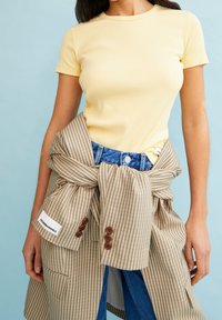 Žluté žebrované tričko spojené s béžovou kostkovanou košilí uvázanou kolem pasu, s hnědými knoflíky a džínami z denimu. Pozadí je světle modré.