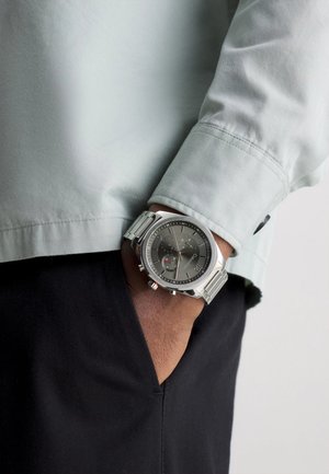 Montre en métal argenté avec un cadran gris, des détails de chronographe et des accents rouges. Portée sur un poignet avec une chemise à manches longues de couleur vert clair.