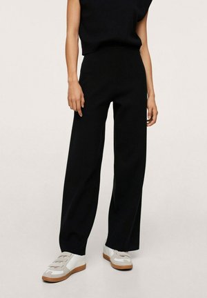 Personne portant un pantalon noir taille haute à jambes larges associé à des baskets blanches avec des accents beiges sur un fond clair uni.