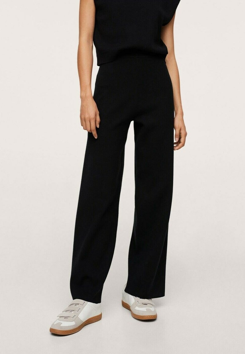 Personne portant un pantalon noir taille haute à jambes larges associé à des baskets blanches avec des accents beiges sur un fond clair uni.