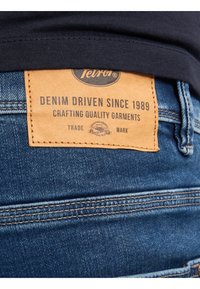 Vaqueros de mezclilla con una etiqueta de cuero marrón que presenta "Impulsados por el Denim Desde 1989" y "Fabricando Prendas de Calidad", con costuras azules y un corte clásico.