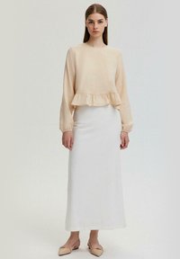 Lange mouw, lichtbeige top met een ruchesrand, gecombineerd met een witte, enkel-lange rok. Het materiaal lijkt zacht en textuur te hebben. Perzikkleurige ballerina's maken de look af.