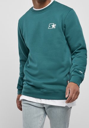 Homme portant un sweat-shirt col ras du cou bleu sarcelle avec un logo étoile sur la poitrine, superposé à une chemise blanche, associé à un jean bleu clair.