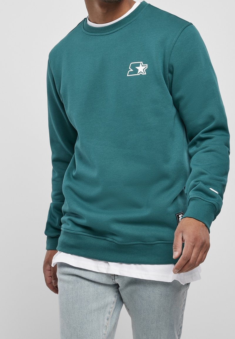 Homme portant un sweat-shirt col ras du cou bleu sarcelle avec un logo étoile sur la poitrine, superposé à une chemise blanche, associé à un jean bleu clair.