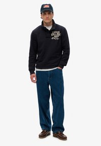 Felpa blu con colletto e testo beige, jeans blu e scarpe marroni. Cappellino con un design della bandiera americana.