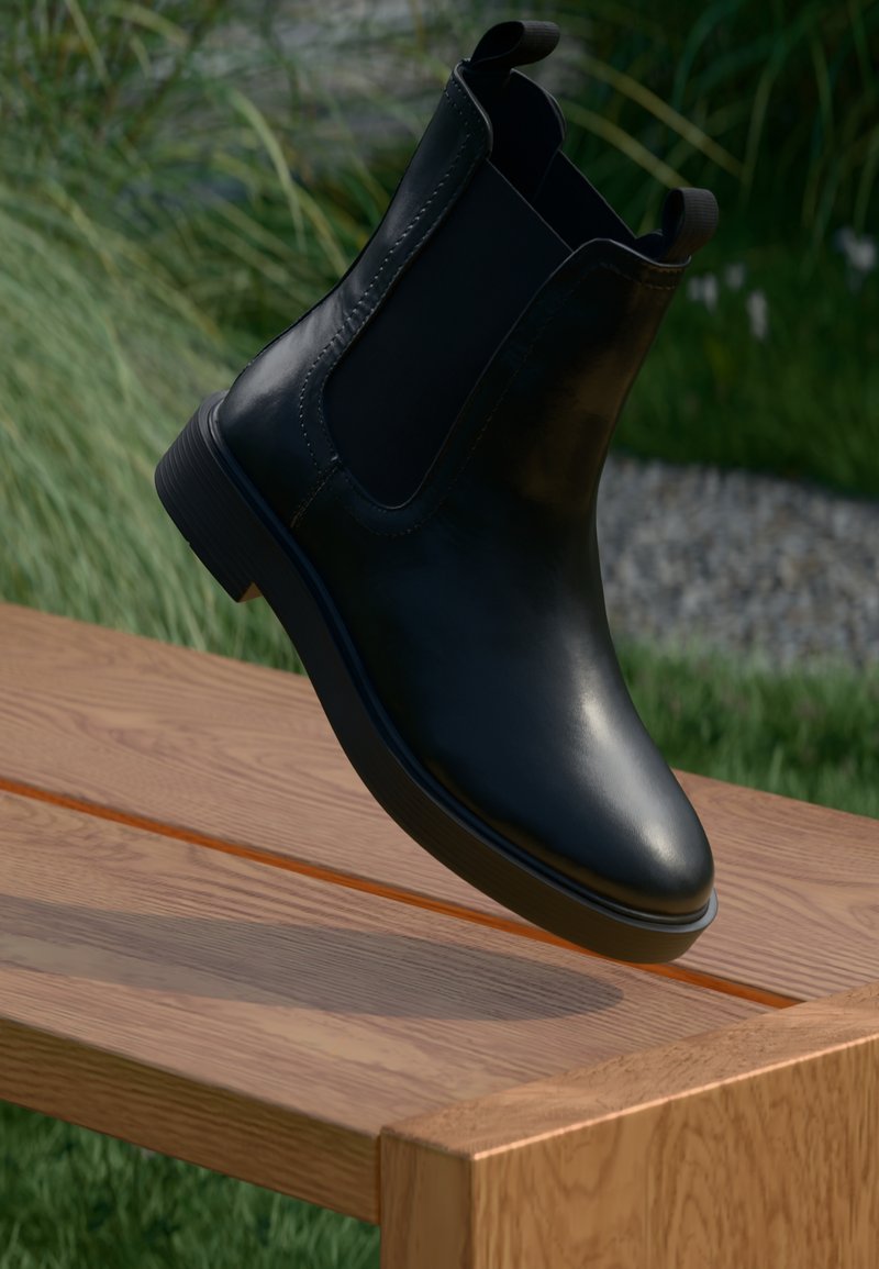 Bota de tornozelo em couro preto com painéis laterais elásticos e salto baixo, apresentando um acabamento liso e bico arredondado, posicionada acima de uma superfície de madeira.