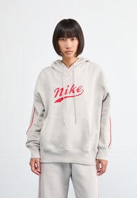 Gaišs pelēks hoody ar sarkano "Nike" logotipu, kabatu priekšpusē, ribotām piedurknēm un sarkanām sānu svītrām. Apvienots ar atbilstošām pelēkām biksēm.