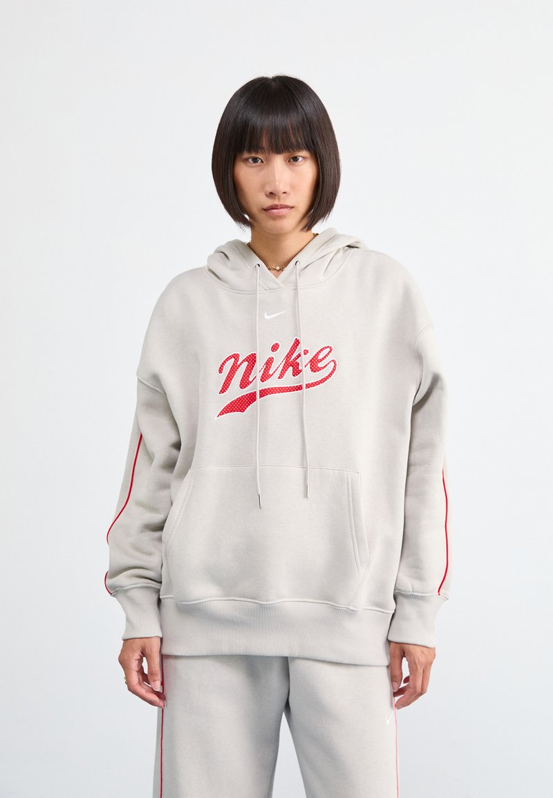 Gaišs pelēks hoody ar sarkano "Nike" logotipu, kabatu priekšpusē, ribotām piedurknēm un sarkanām sānu svītrām. Apvienots ar atbilstošām pelēkām biksēm.