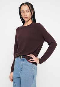 Pulover tricotat burgundy cu mâneci lungi, decolteu rotund și croială lejeră, asortat cu blugi cu talie înaltă, de culoare albastru deschis. Textură netedă.