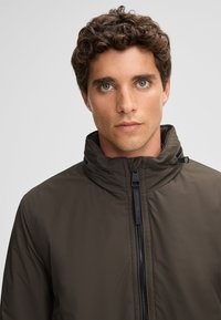 Braune Zip-Jacke mit hohem Kragen, glatter Textur und subtiler Ausstrahlung; verfügt über einen Frontreißverschluss und Markendetails entlang des Reißverschlusses.