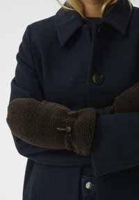 Námornícky vlnový kabát s čiernymi gombíkmi, nosený cez tmavý sveter. Hnedé fleecové rukavice s detailom na šnúrku zakrývajú ruky.