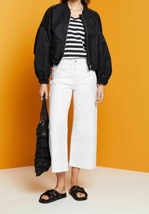 Giacca bomber nera con maniche oversize, maglietta a righe bianche e nere, jeans bianchi a gamba larga, sandali neri scivolati e borsa nera texturizzata.