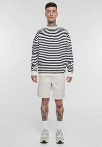 Stripete langermet skjorte i svart og hvitt, kombinert med lyse beige shorts. Modell iført lyse joggesko, hvite sokker, og viser frem tatoveringer.