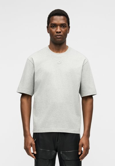 Νέος άνδρας στέκεται με πρόσωπο προς τα εμπρός φορώντας ένα ανοιχτό γκρι κοντομάνικο μπλουζάκι και μαύρο παντελόνι cargo με μπροστινές τσέπες.