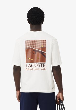 Hombre con camiseta blanca con un gráfico de cancha de tenis, pelotas de tenis y el texto "LACOSTE Roland Garros Club" en la parte trasera.