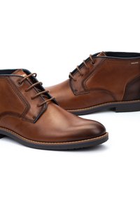 Botas de tobillo de cuero marrón con textura suave, punta redondeada y diseño con cordones. Incluye acentos oscuros contrastantes y suela de goma duradera.