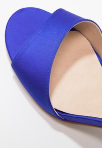 Modré saténové skluzné sandály s otevřenou špičkou a širokým popruhem s bé�žovou polstrovanou stélkou na bílém pozadí.
