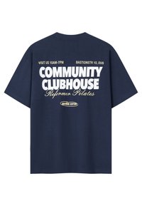 Marineblå T-shirt med hvid og beige tekst: "Besøg os 10-19 Bastionstr 10, DUS Community Clubhouse Reformer Pilates Another Cotton."