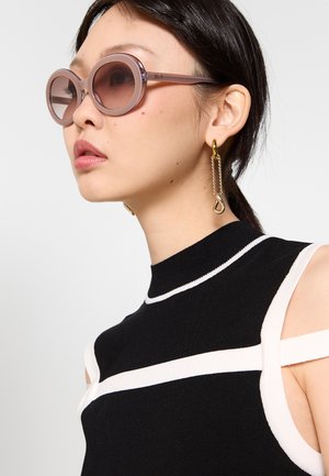 Donna che indossa occhiali da sole rotondi rosa, orecchini a catena dorata pendenti e top nero senza maniche con bordo bianco e dettaglio cut-out sulla spalla.