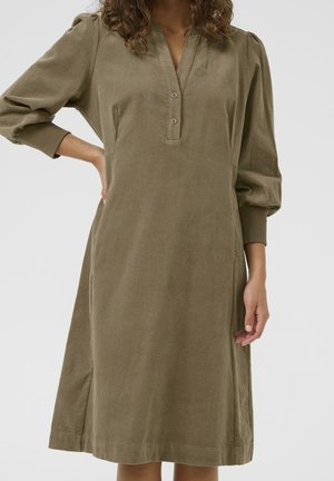 Kobieta ubrana w oliwkową, długorękawową sukienkę sztruksową z dwukolumnowym zapięciem na guziki i ściągaczami na rękawach, stojąca z jedną ręką na biodrze.