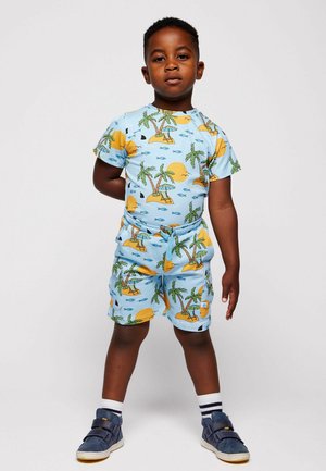 Noor poiss seisab, kandes sinakaid troopiliste palmipuude, päikese, kaladega sobivaid t-särki ja lühikesi pükse ning tumesiniseid saapaid.