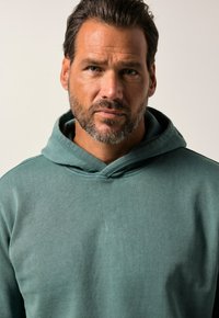 Sudadera verde claro hecha de tela suave, con capucha ajustable y un corte relajado. Sin patrones ni acentos visibles. Diseño sencillo.